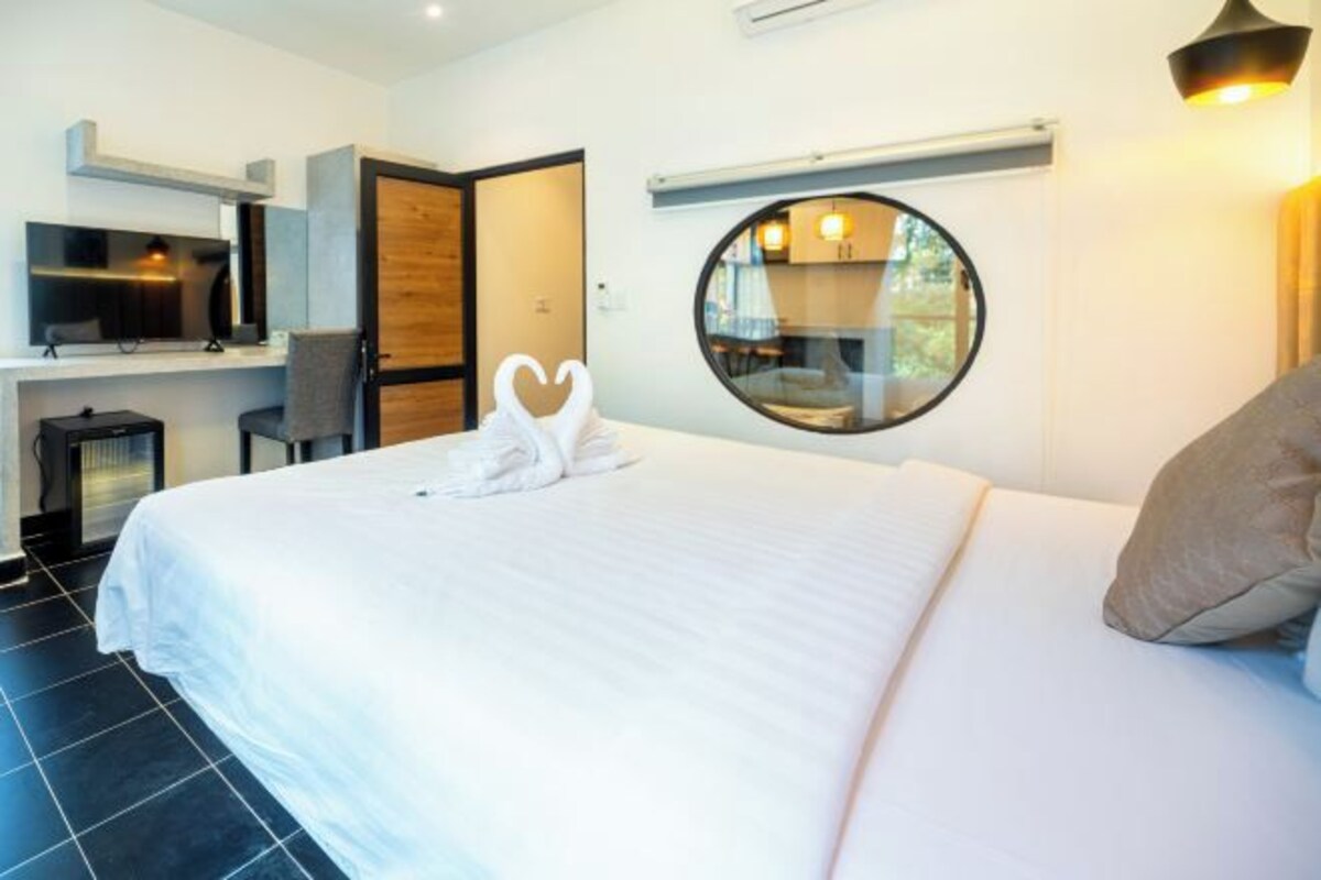 Ein modernes Hotelzimmer mit einem großen Bett, einem Schreibtisch mit Flachbildfernseher, einem runden Spiegel und Blick ins Freie durch ein Fenster.