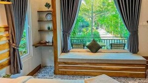 A room with a window, curtains, a bed, a small table, and a shelf with decorative items. - Raing Phnom Bungalow (Kampot)