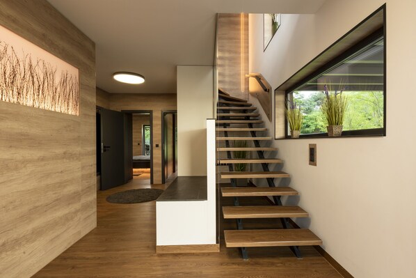 A modern staircase with wooden steps and a glass railing, leading to a landing with a large window overlooking greenery.