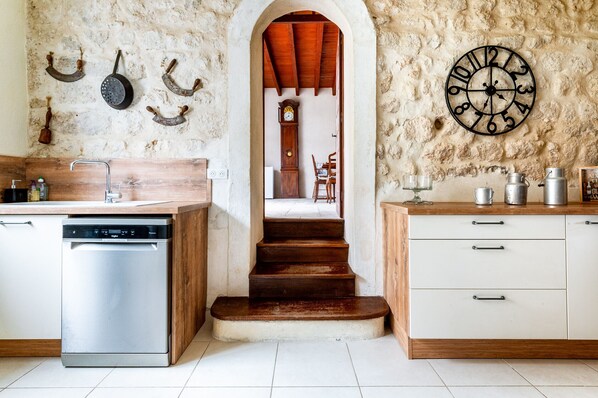A kitchen with a dishwasher, wooden cabinets, a clock, and a rustic stone wall. - House in listed village with garden and jacuzzi (La Garde-Adhémar)