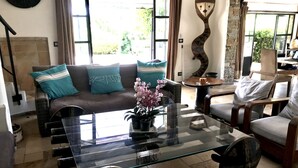 A living room with a glass coffee table, a sofa with teal cushions, a decorative wall piece, and a view through large windows. - Charming residence in Grimaud (Grimaud)
