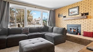 Una sala de estar con sofá modular, reposapiés, chimenea y ventanas amplias.