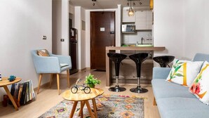 Una sala moderna con sofá, mesa de centro y comedor con una silla y una mesita. Se observa un área de cocina con microondas y pileta.