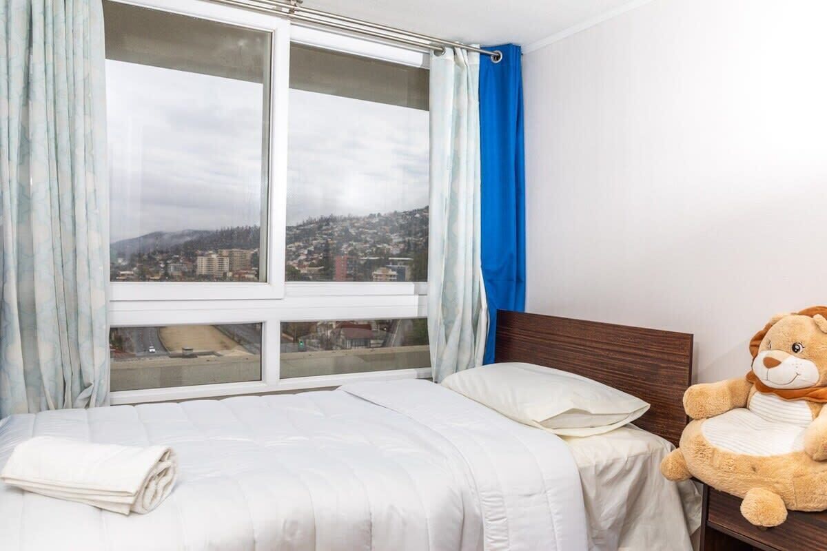 Une chambre d’hôtel avec un lit, une fenêtre donnant sur un paysage urbain et un jouet en peluche posé sur la table de chevet.