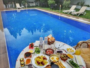 En frukostservering vid poolkanten med ett bord fullt av mat och dryck, omgiven av poolen och solstolar.