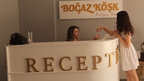 Una mesa de recepción con la palabra "RECEPTİ" en letras grandes, un letrero encima que dice "BOGAZ KÖŞK Boutique Hotel", y una mujer de pie junto al mostrador.