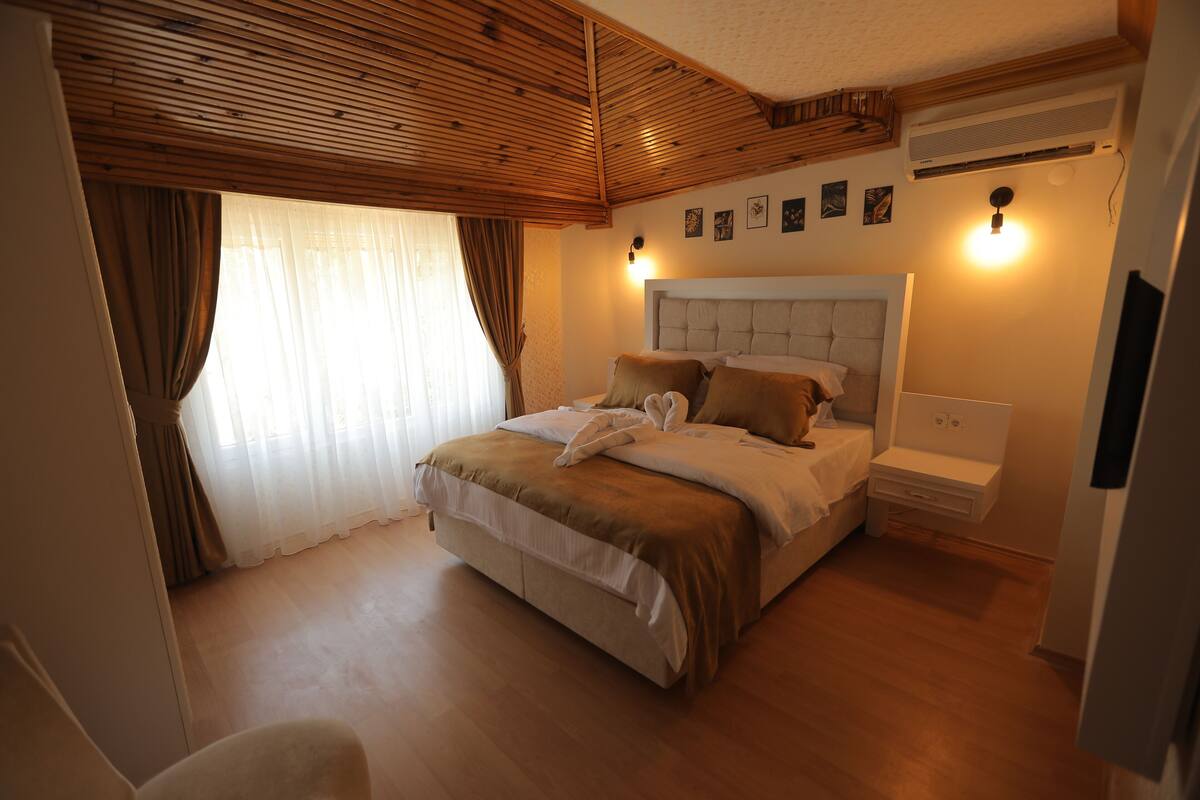A bedroom with a wooden ceiling, a bed with a headboard, a bedside table, and a wall-mounted air conditioner.