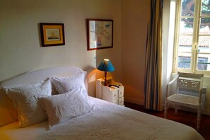 Un letto rifatto con lenzuola bianche e dettagli in pizzo, una lampada da comodino, una sedia e due quadri appesi al muro.