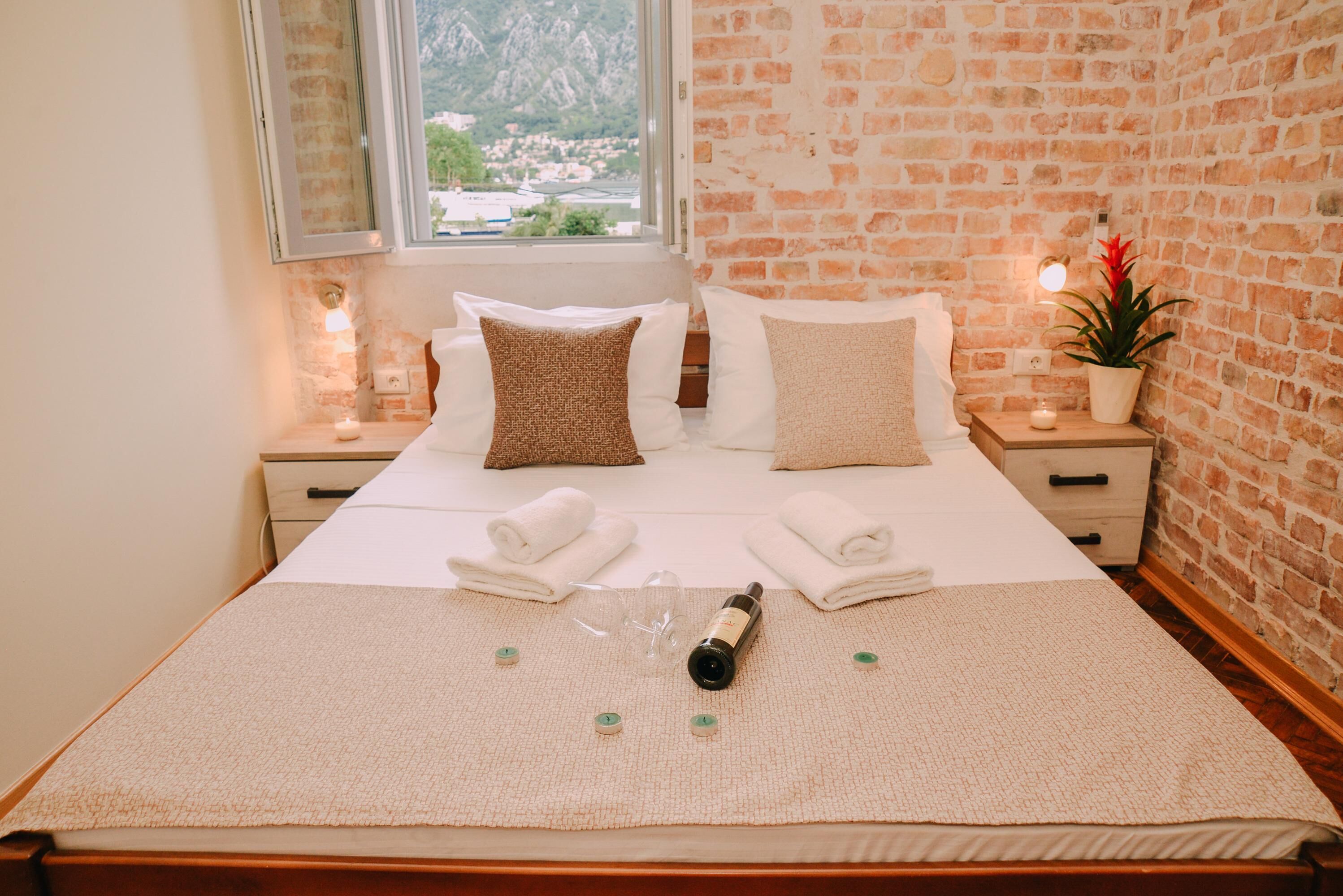 A neatly made bed with pillows, a bottle, and glasses on a textured bedspread. There are two bedside tables with lamps and a potted plant. A window with a view of mountains and a village is visible.