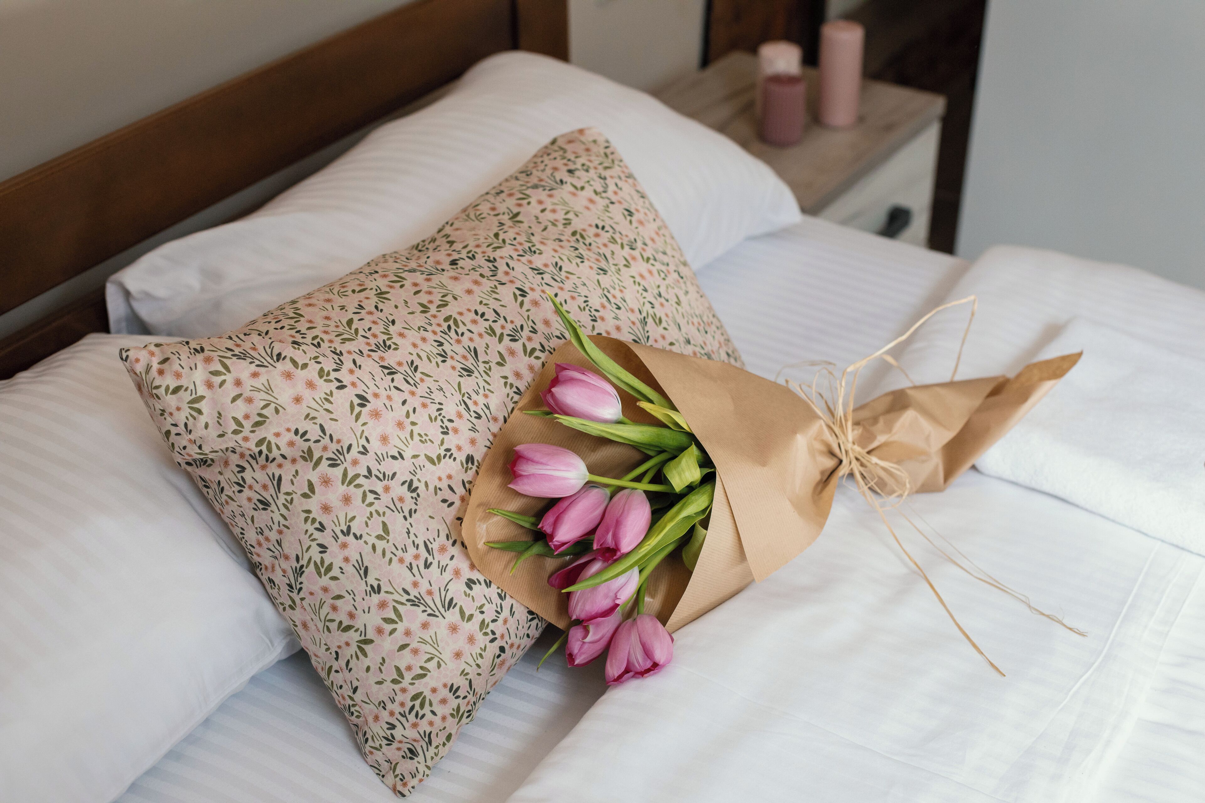 A bed with white linens, a floral pillow, and a bouquet of pink tulips tied with twine.