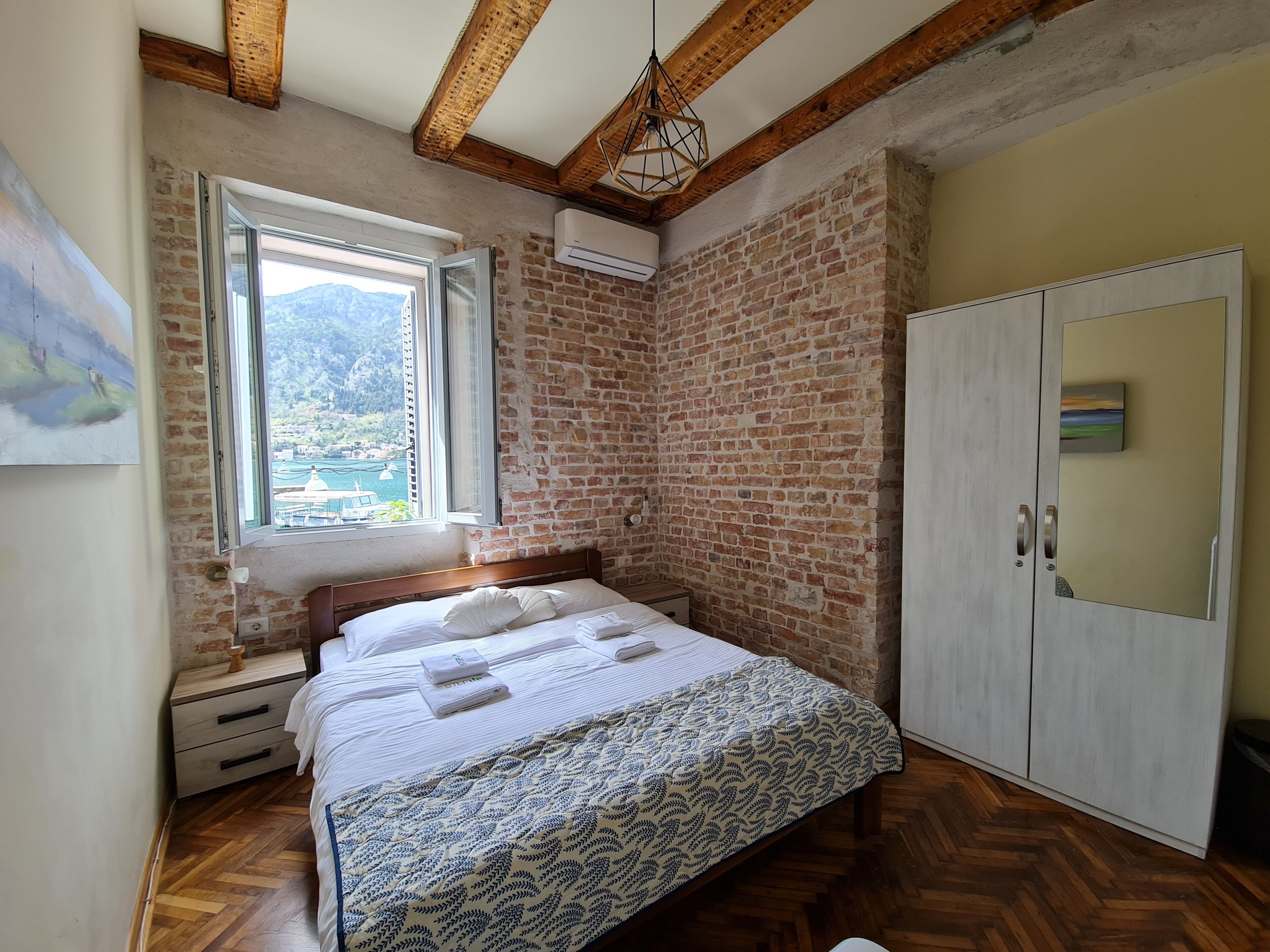 A bedroom with a brick wall, wooden floor, a bed with a patterned bedspread, a nightstand, a wardrobe, and a window with a view of a body of water and mountains.