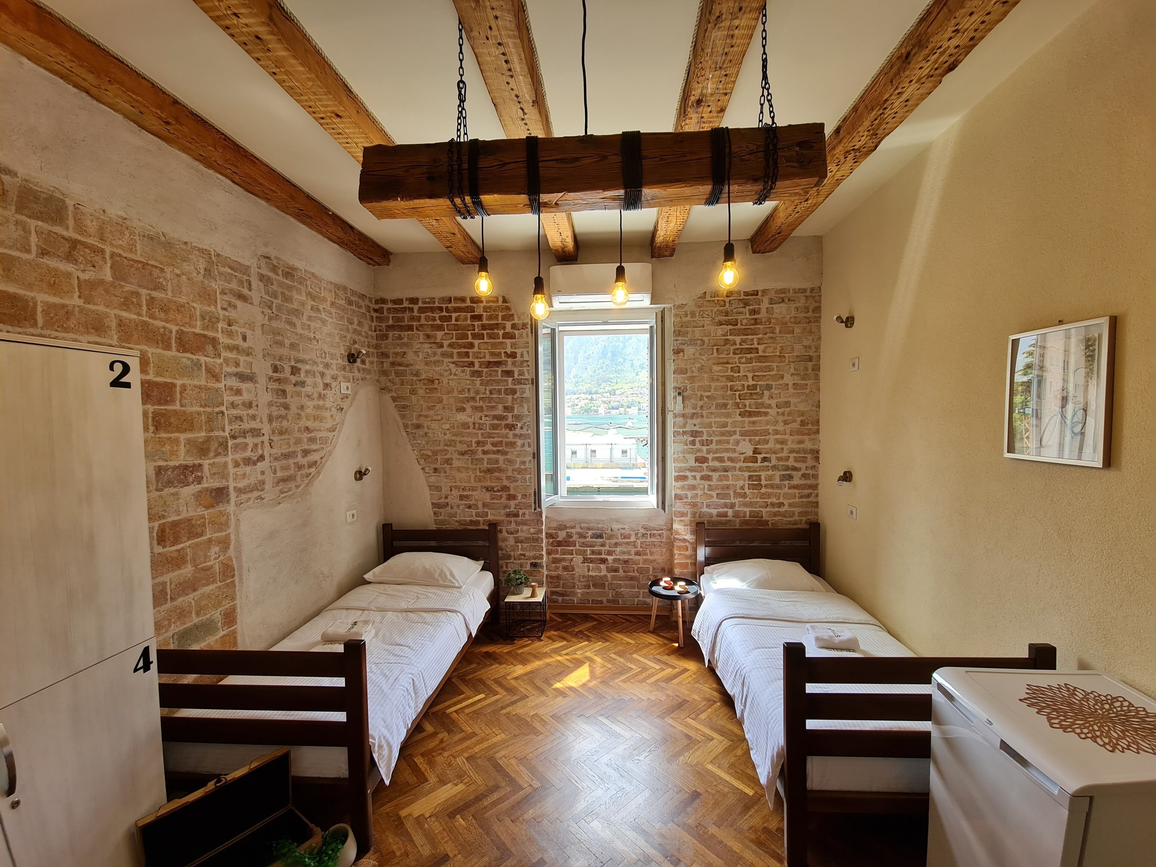 A room with two beds, wooden beams, brick walls, and a window with a view.