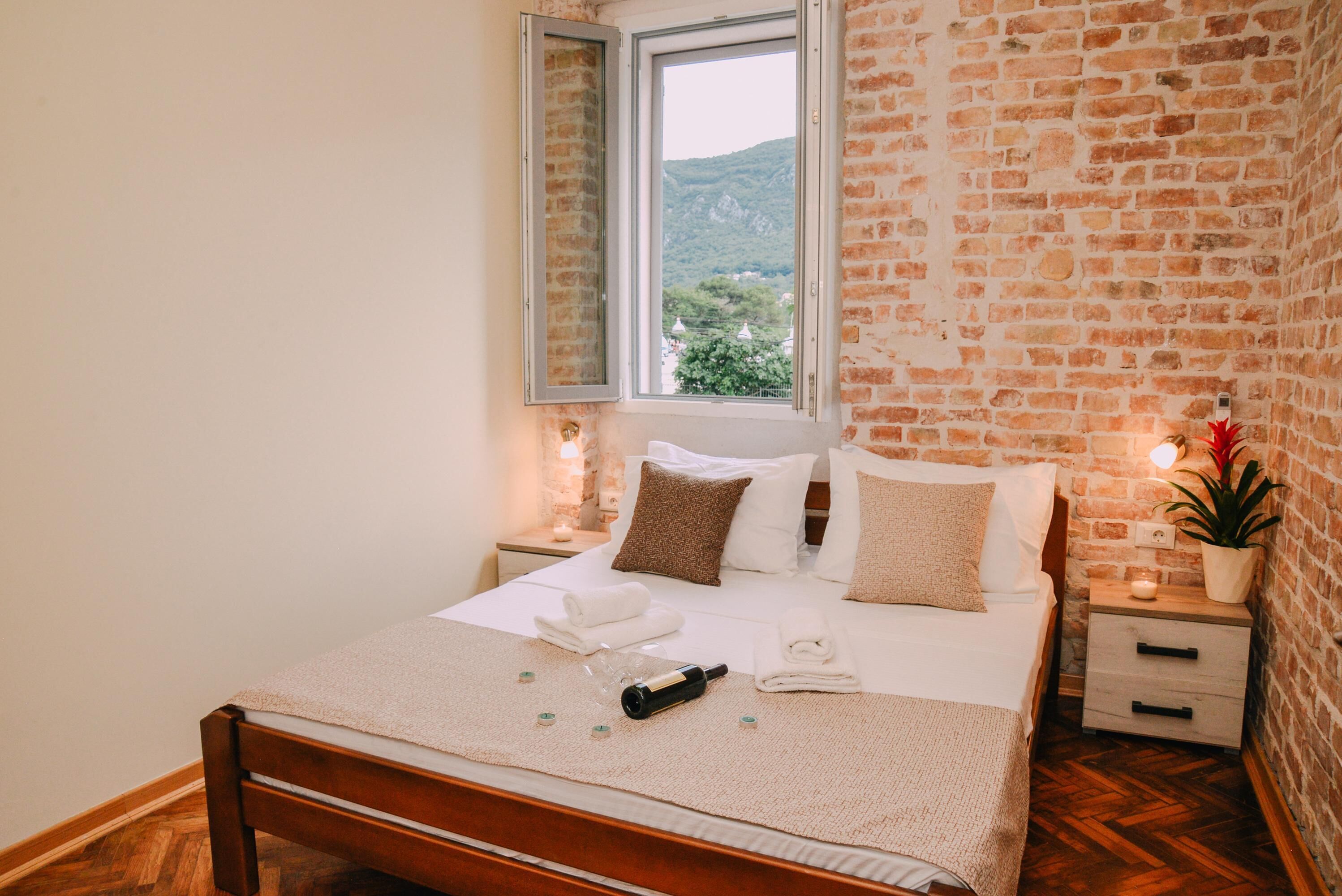 Una cama bien hecha con cabecera de madera, una mesita de noche con lámpara y vista a la montaña a través de una ventana.