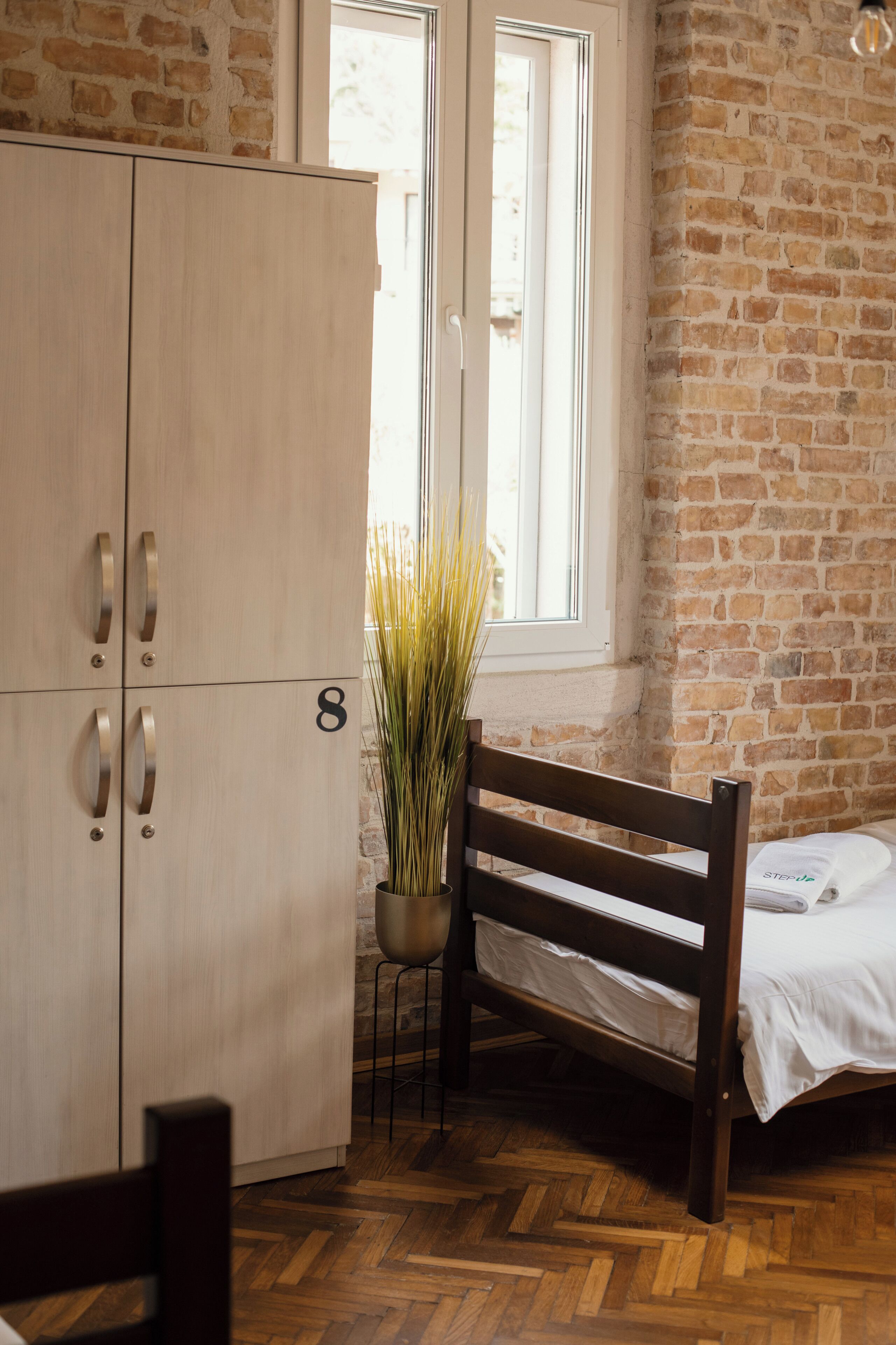Una habitación con una cama de madera, una silla de madera, un armario de madera, una planta en maceta y una ventana con vistas al exterior.