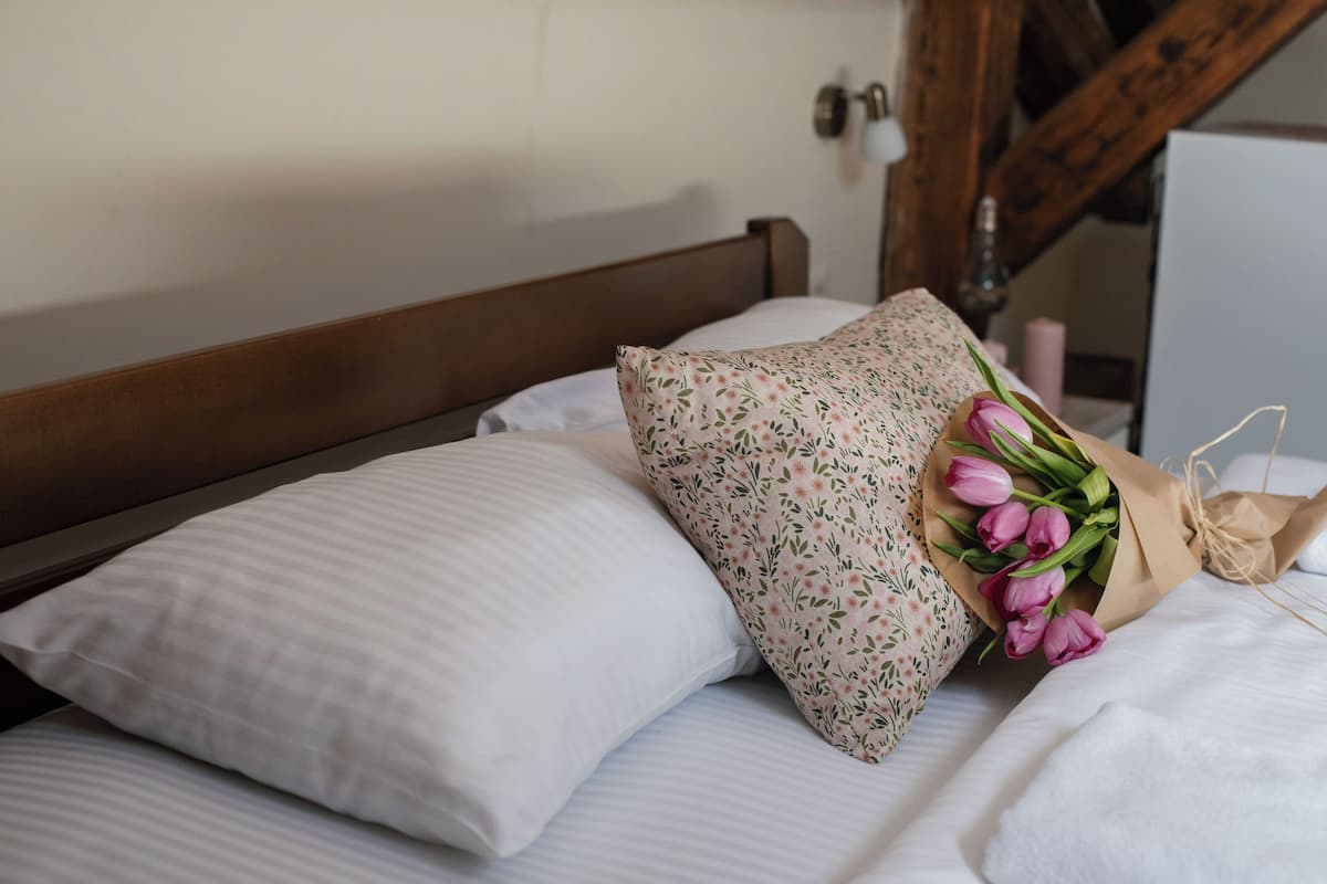 En säng med vita sängkläder och en prydnadskudde med blommönster, en bukett med rosa tulpaner och en sänggavel i trä.