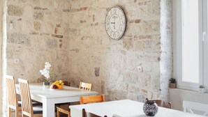 Un comedor con pared de piedra, sillas de madera, una mesa blanca y un reloj de pared.