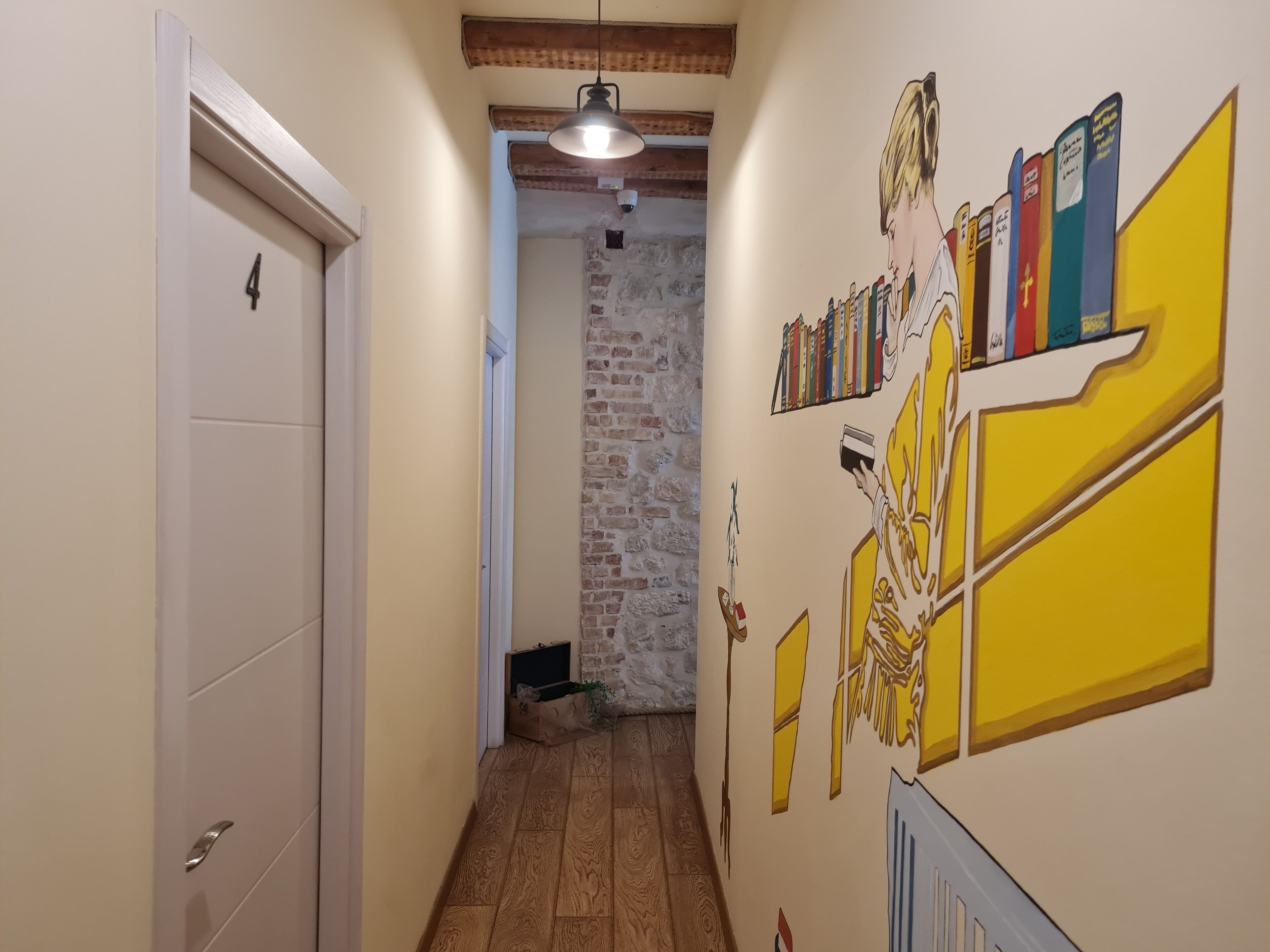 Un passadís amb una porta blanca marcada amb el número 4, una paret de pedra i un mural amb llibres i una figura.