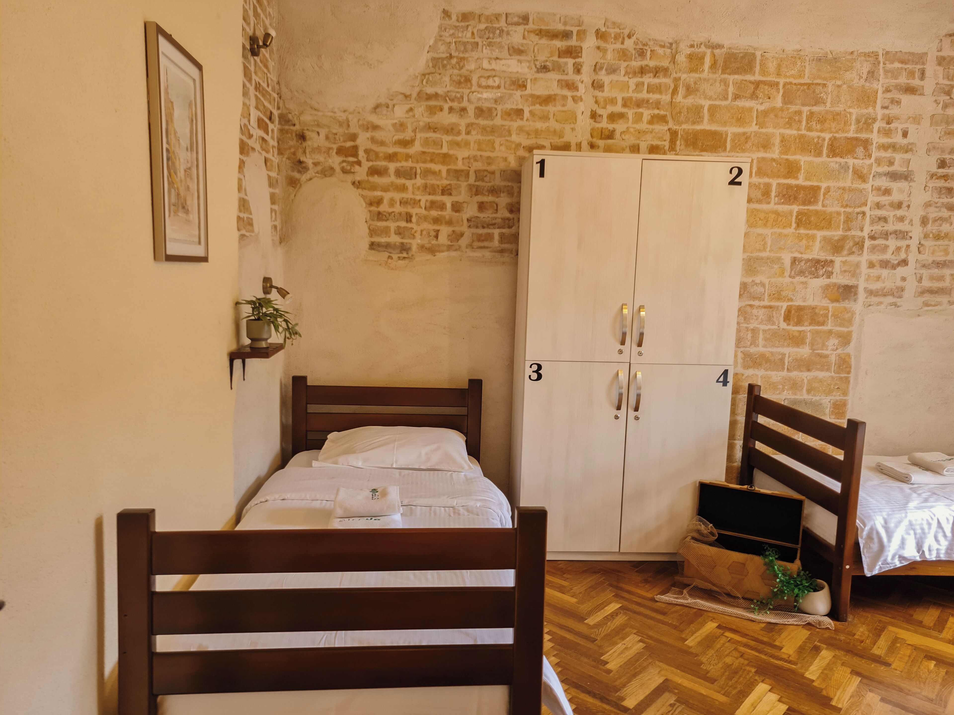 A room with a wooden bed, a wardrobe with four doors, a small shelf with a plant, and a herringbone-patterned wooden floor.