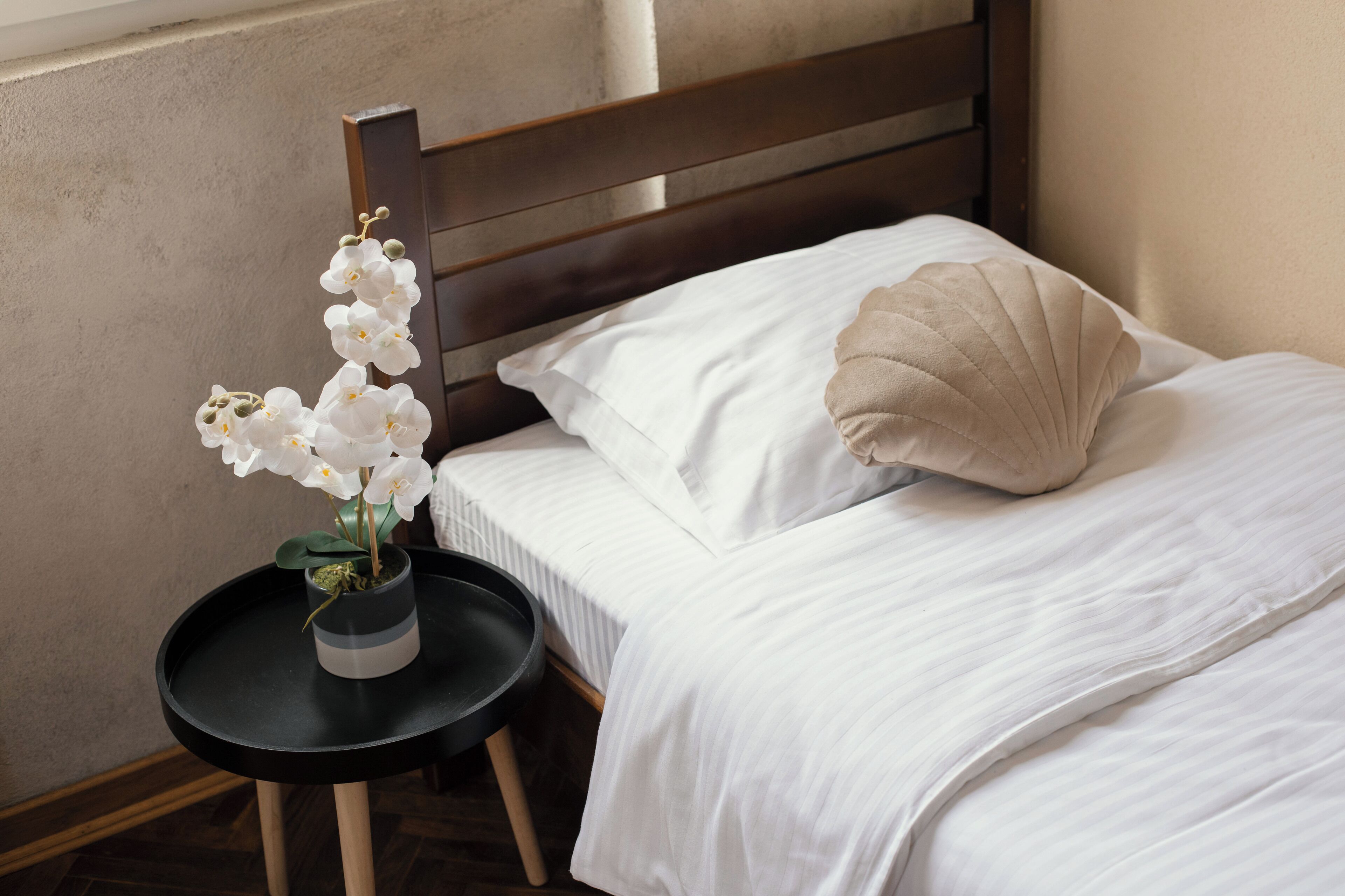 A neatly made bed with white bedding and a beige pillow, a small black table with a vase of white flowers, and a wooden headboard.