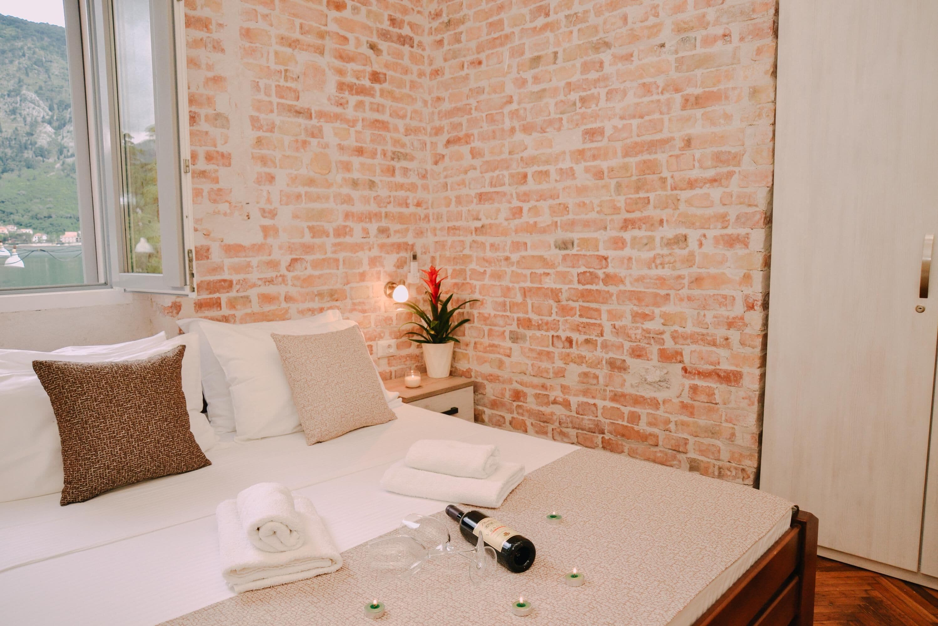 Une chambre d’hôtel avec un mur en briques, un lit recouvert de draps blancs, une table de chevet avec une lampe et une plante, et une fenêtre donnant sur un paysage verdoyant.