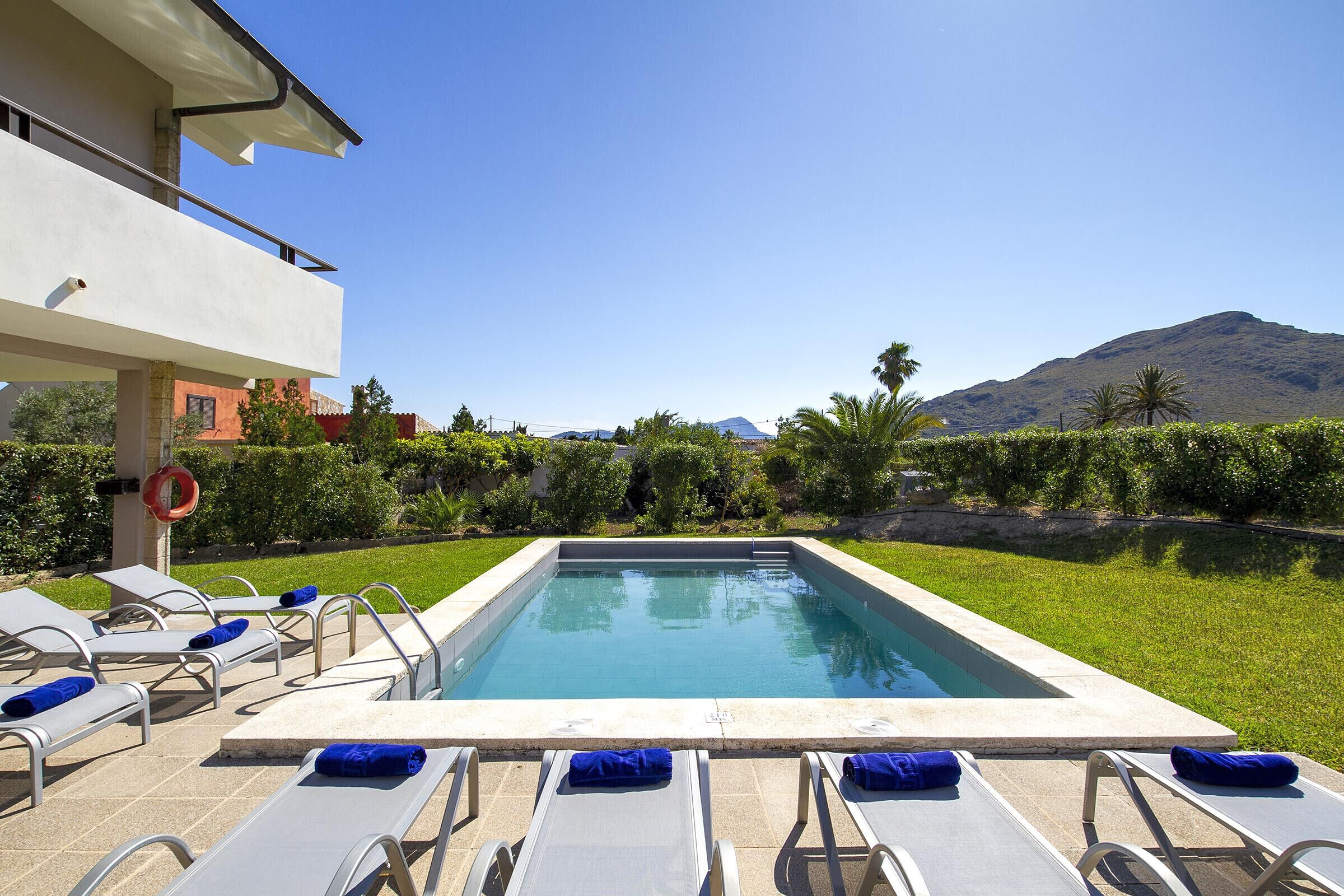 A rectangular swimming pool with lounge chairs and a lifebuoy, surrounded by greenery and a mountain in the background.