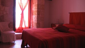 A bedroom with a stone wall, a bed with a red bedspread, a chair, and a window with sheer curtains. - JATTA - Beautiful Stone House Puglia (Conversano)