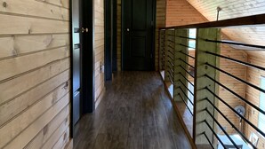Un couloir intérieur en bois, avec un plancher en bois foncé, une porte noire et une rambarde en verre sur le côté droit.