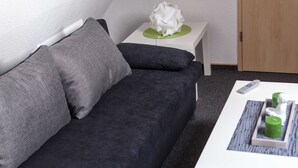 Un sofá gris oscuro con dos almohadas, una mesita de noche blanca con un objeto blanco, y una mesa de centro blanca con un control remoto y un objeto verde.