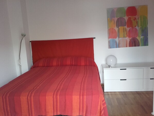 A room with a red-striped bed, a white bedside table with drawers, and a colorful abstract painting on the wall.