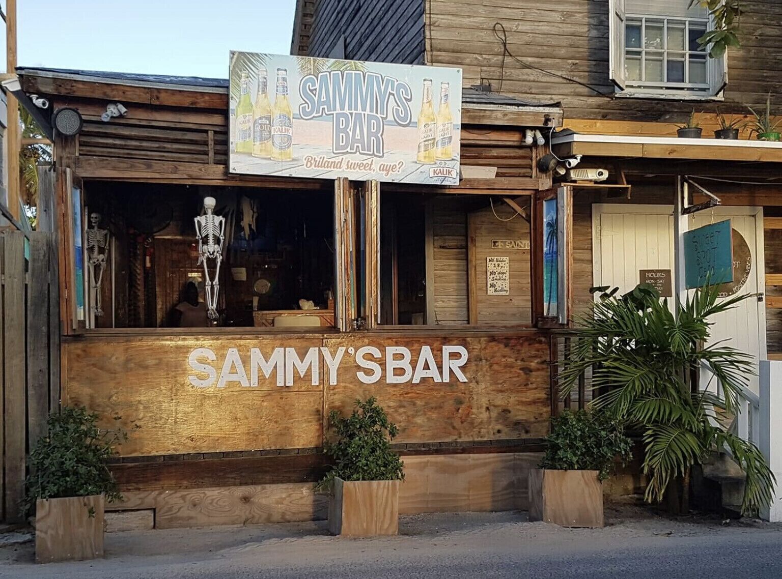 Un bar de madera rústico llamado Sammy's Bar, con una decoración de esqueleto, contraventanas de madera y plantas en macetas.