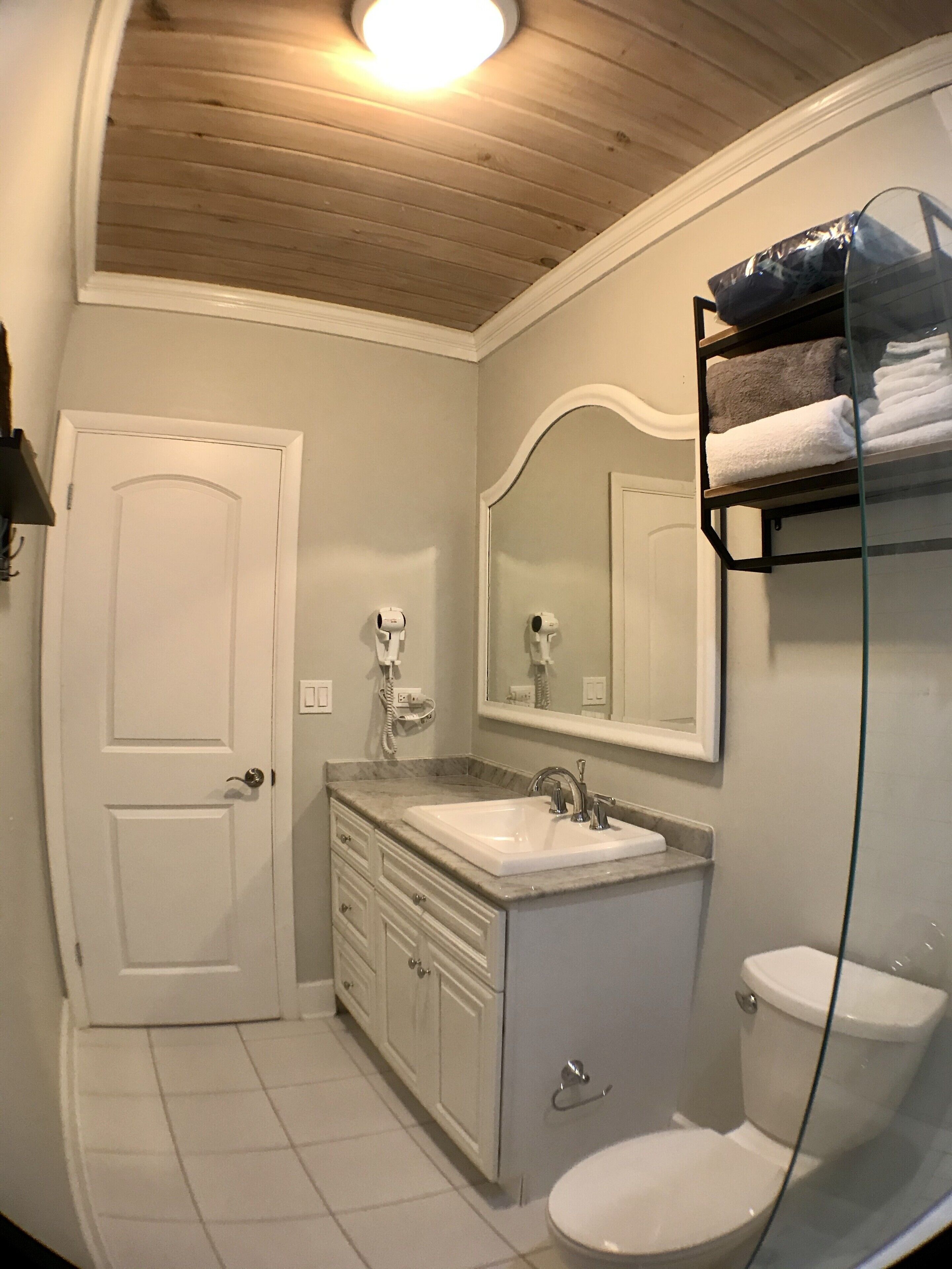 Un baño con lavamanos, tocador y retrete blancos. Hay un espejo sobre el lavamanos y una estantería con toallas.