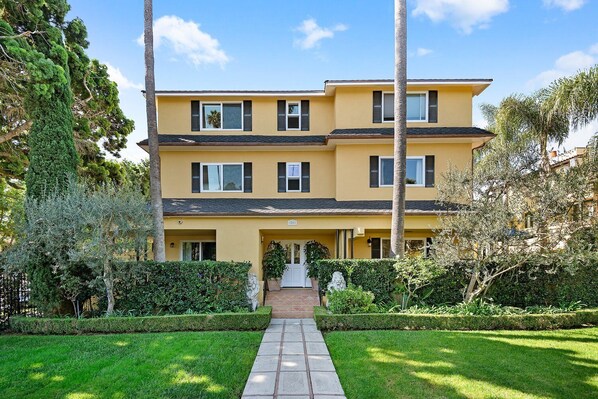 A two-story yellow building with a front entrance, surrounded by well-maintained greenery and a stone pathway leading to the door. - Historic West Beach Studio — blocks to beach and Funk Zone (Santa Barbara)