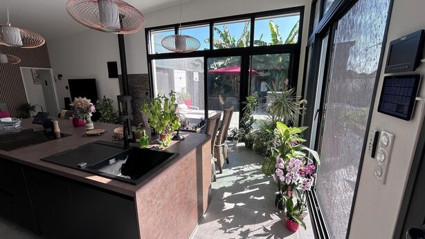 A modern kitchen with a large island, a dining area with wicker chairs, and a view of the outdoors through large windows. - New house, Jacuzzi, Terrace, Near to Beaches and Shops (Le Tour-du-Parc)
