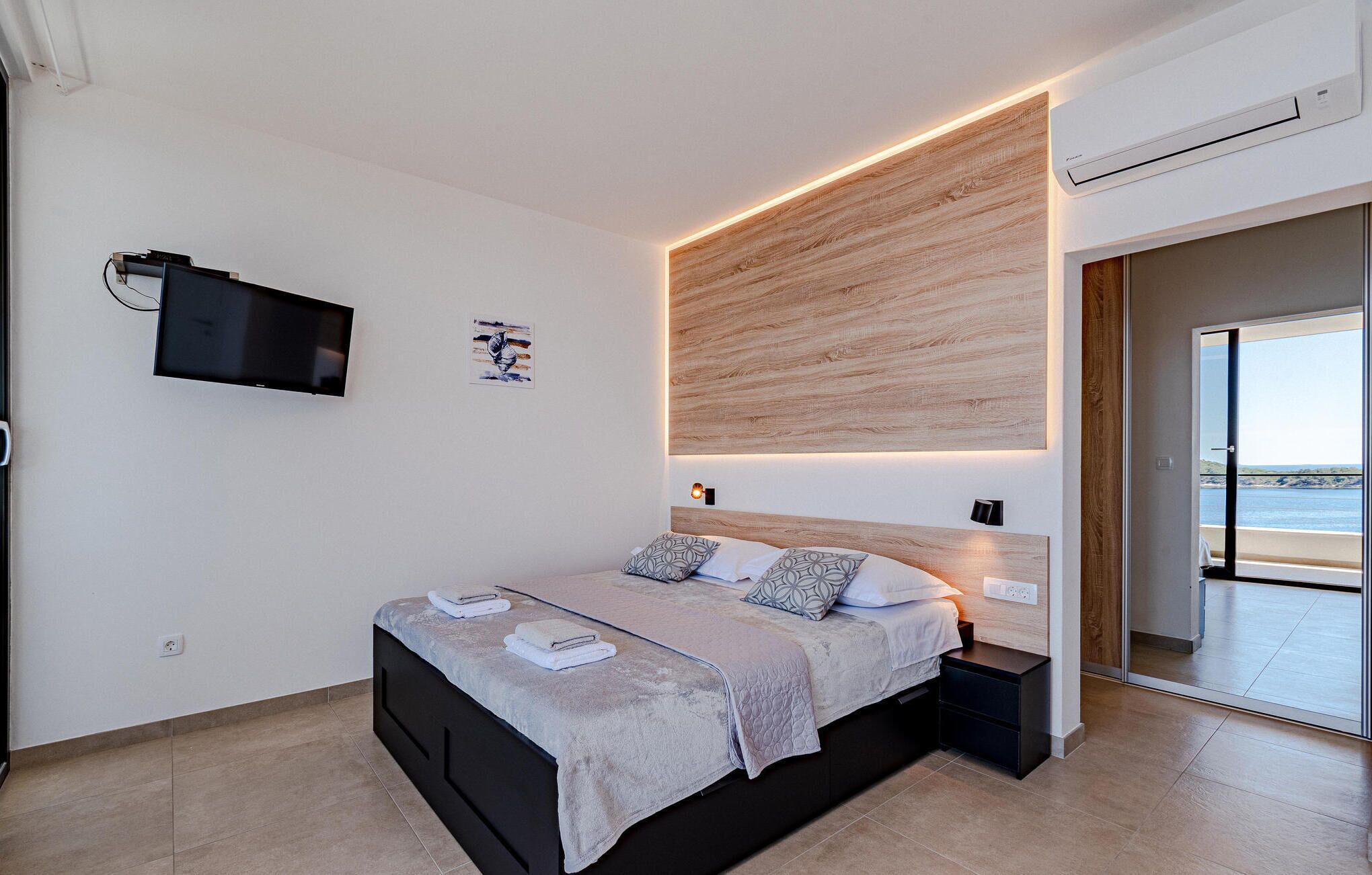 A modern bedroom with a bed, bedside tables, a TV, and a view of the sea.
