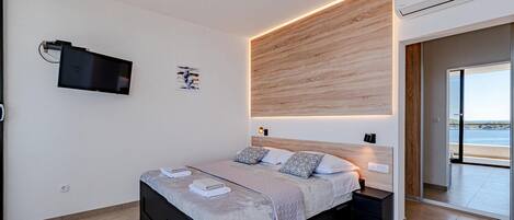 A modern bedroom with a bed, bedside tables, a TV, and a view of the sea.