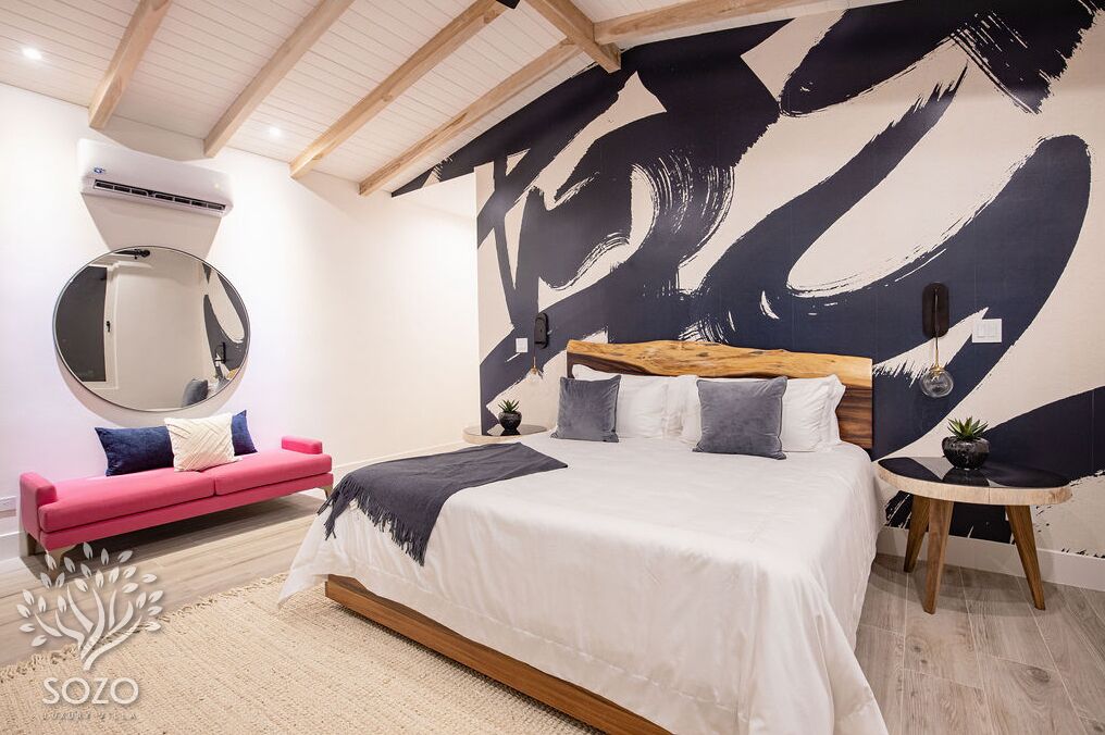 Quarto com uma cama grande, um sofá rosa, um espelho redondo e uma parede com um design abstrato em preto e branco.