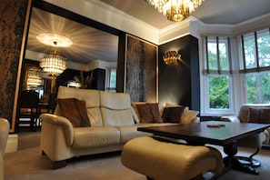 A room with a sofa, ottoman, and a table. There is a chandelier, a mirror, and a window with blinds.