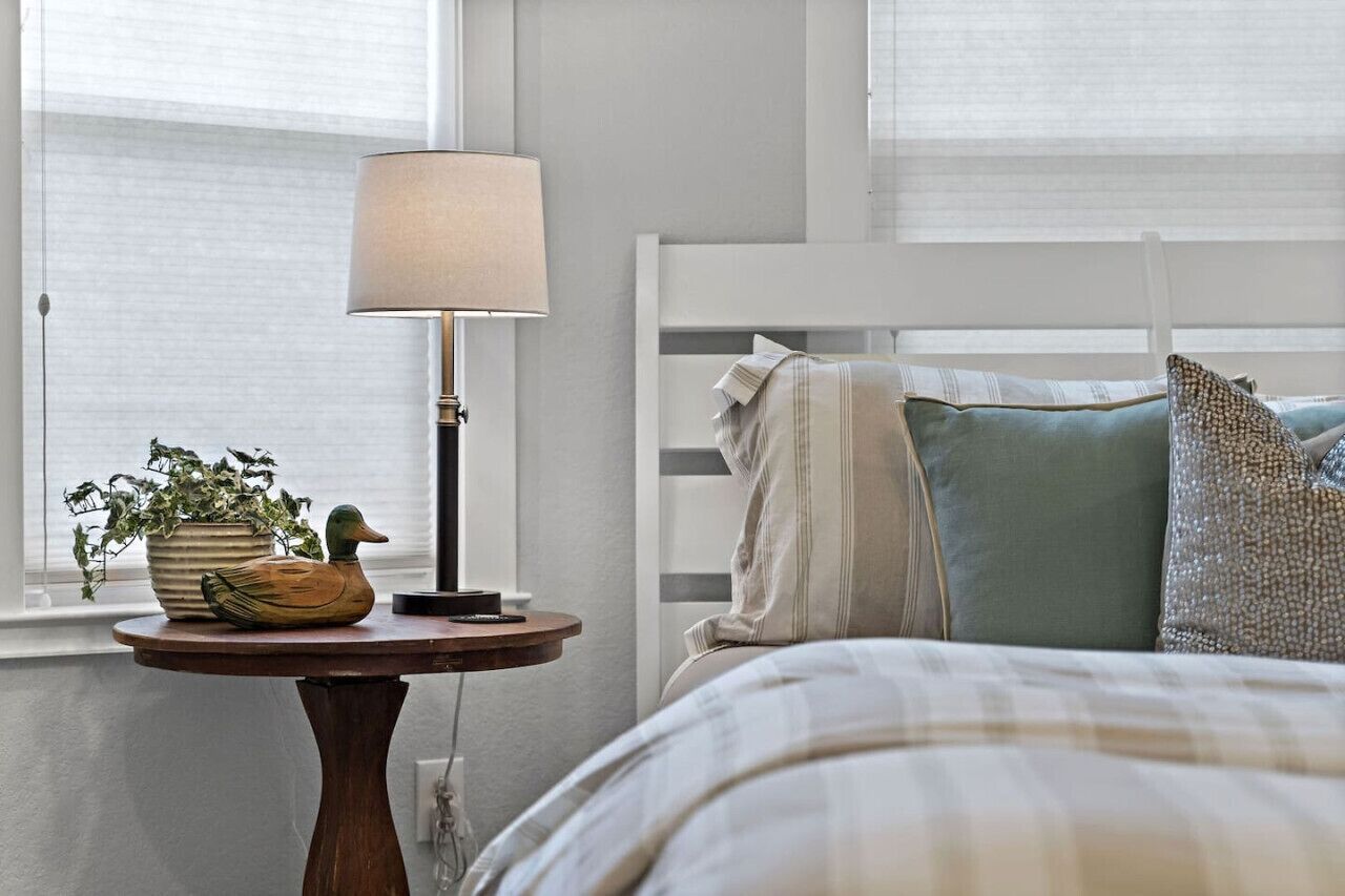 Une chambre à coucher avec un lit, une table de chevet avec une lampe, une plante en pot et une fenêtre avec des stores.