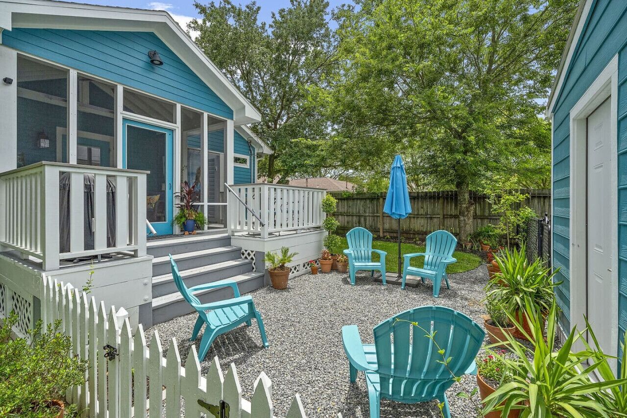 Un patio couvert agrémenté d’une clôture blanche en bois, une véranda avec une porte bleue et plusieurs chaises Adirondack bleues.