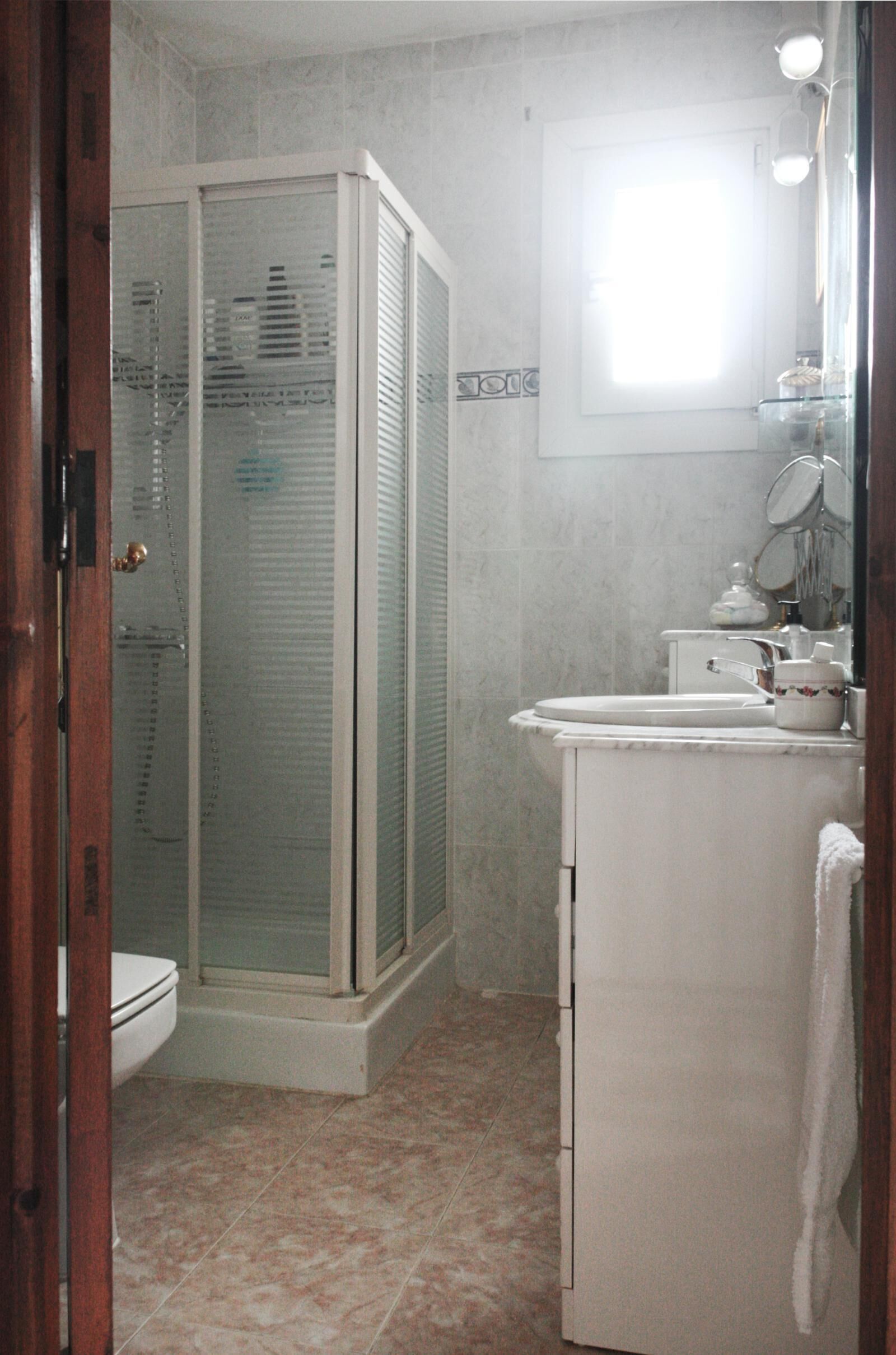 A bathroom with a glass-enclosed shower, a toilet, a sink with a mirror, and a window providing natural light.