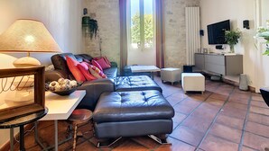 A cozy living room with a sofa, ottoman, and a TV mounted on the wall. - Le 115, air-conditioned town house with interior courtyard and swimming pool (Pernes-les-Fontaines)
