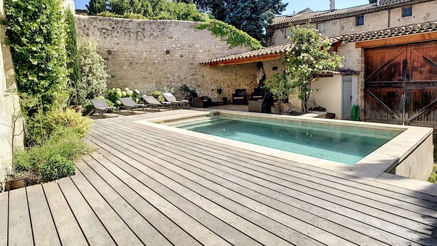 A pool area with a stone wall, wooden deck, and lounge chairs. - Le 115, air-conditioned town house with interior courtyard and swimming pool (Pernes-les-Fontaines)