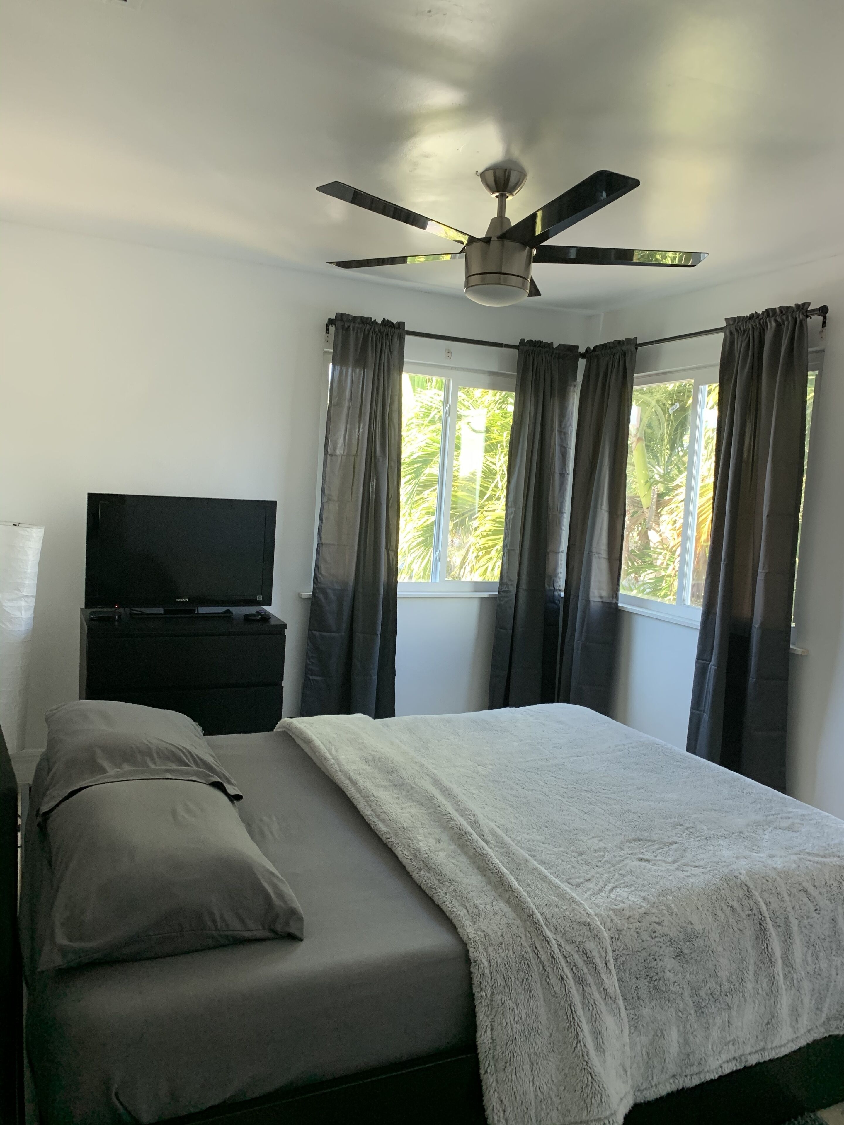 A bedroom with a bed, a TV on a stand, a ceiling fan, and a window with curtains.