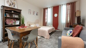 Un dormitorio con cama, sofá, televisor, escritorio con sillas, estantería y ventana con cortinas.