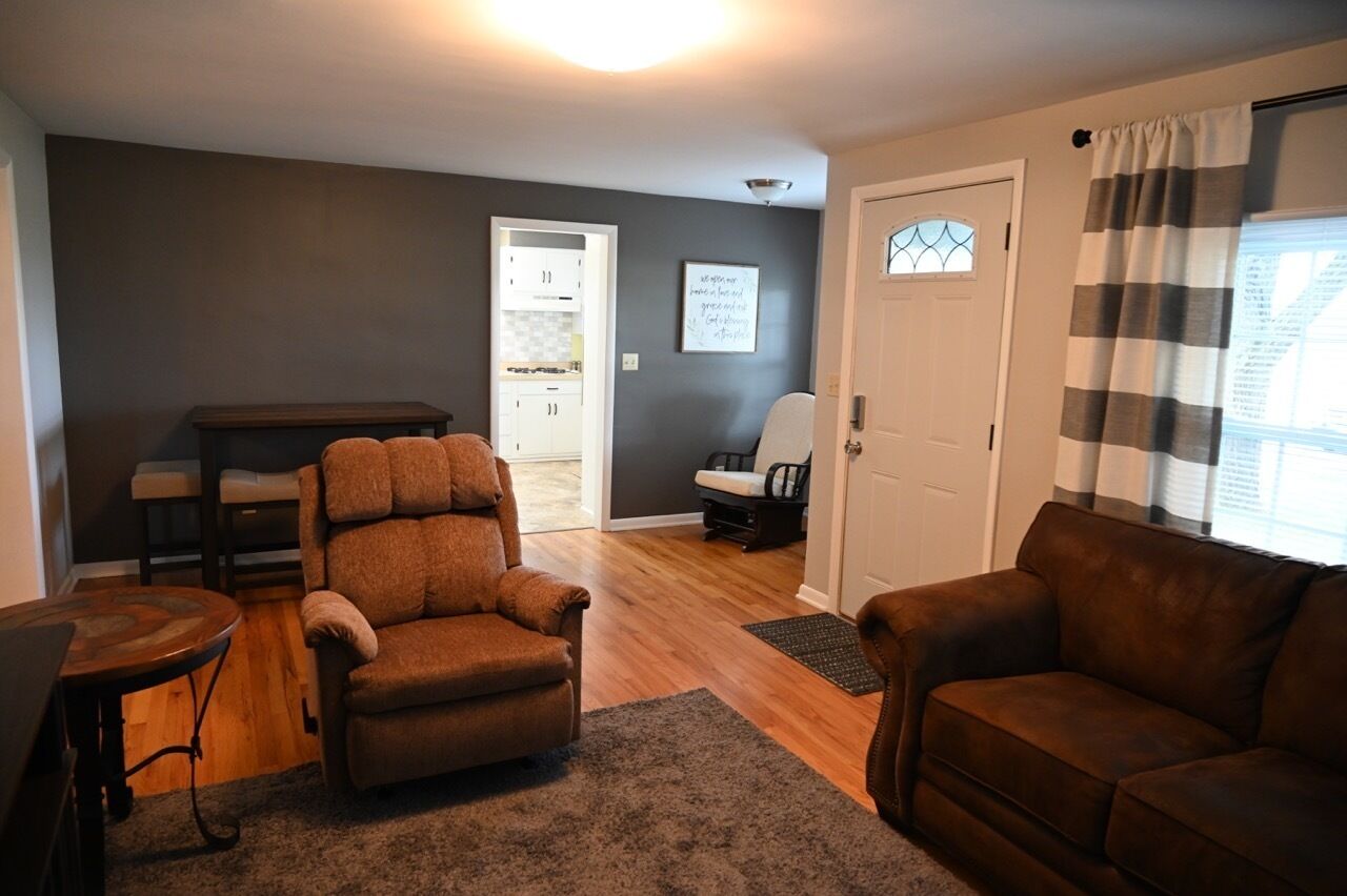 A living room with a brown leather sofa, a wooden coffee table, and a dark rug. There is a door leading to a kitchen and a dining area.