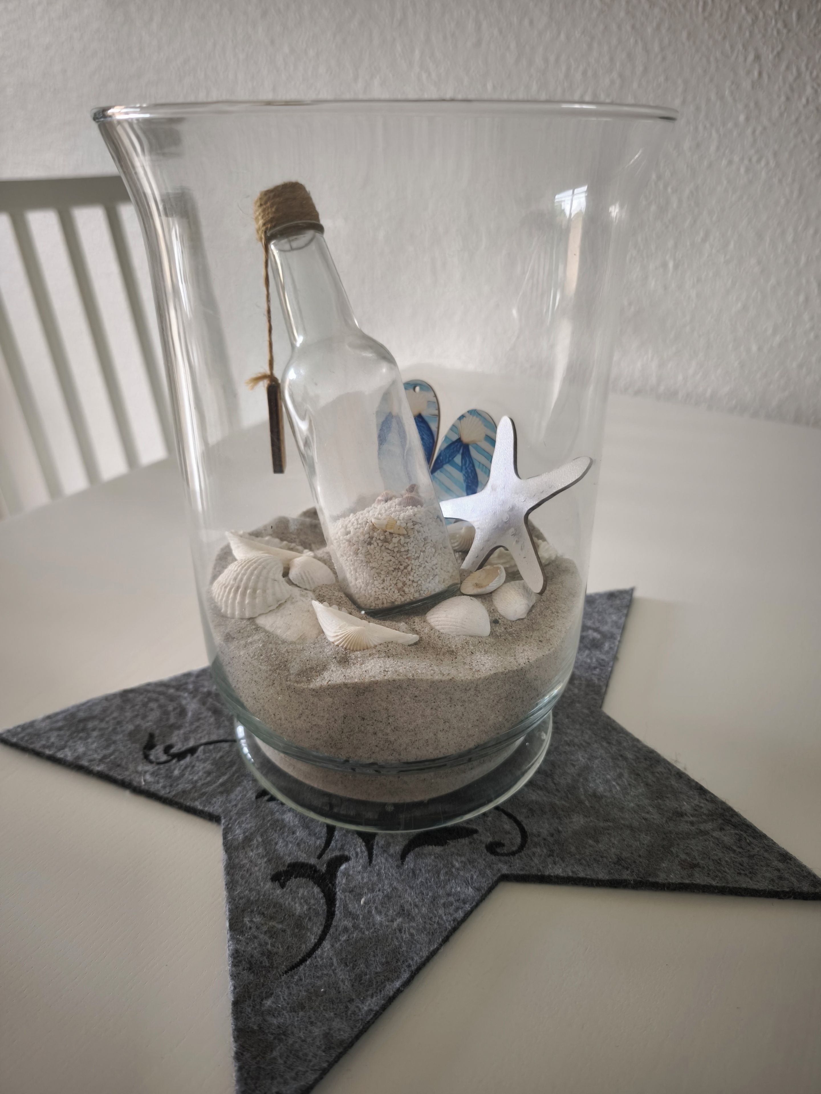 Un bol en verre transparent, muni d’un bouchon en liège, rempli de sable et de coquillages décoratifs, posé sur un dessous de verre en feutre foncé.