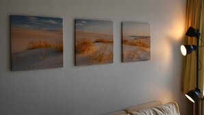 A room with three paintings of desert landscapes on the wall, a bed with a beige cover, and a lamp on the right side.