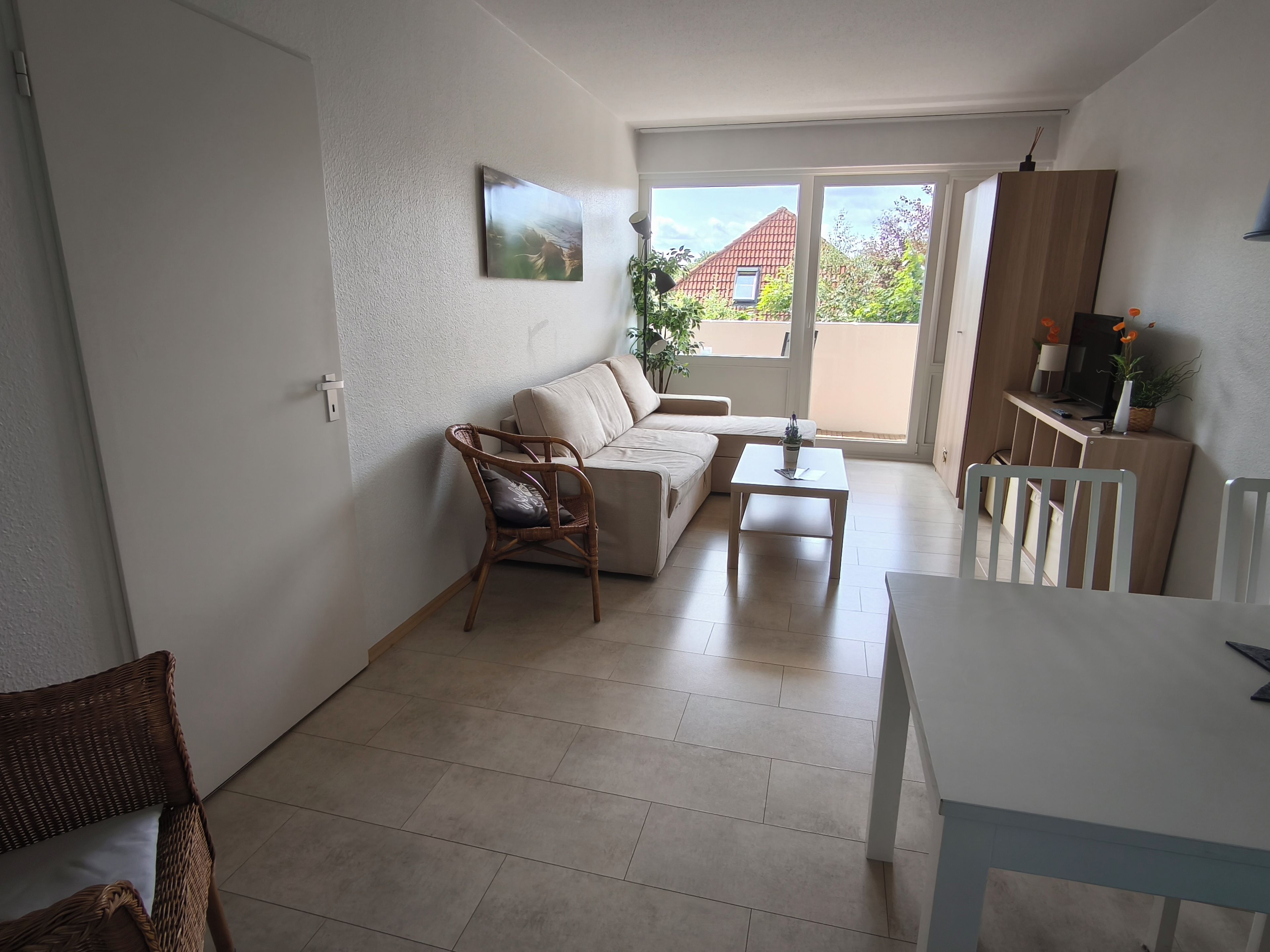 Ein Wohnzimmer mit einer Couch, einem Esstisch und einem Sessel. Ein Fenster bietet Blick auf ein Haus und Grünflächen.