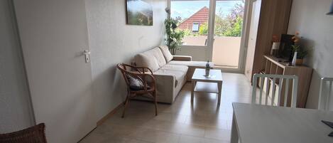 Ein Wohnzimmer mit einer Couch, einem Esstisch und einem Sessel. Ein Fenster bietet Blick auf ein Haus und Grünflächen.