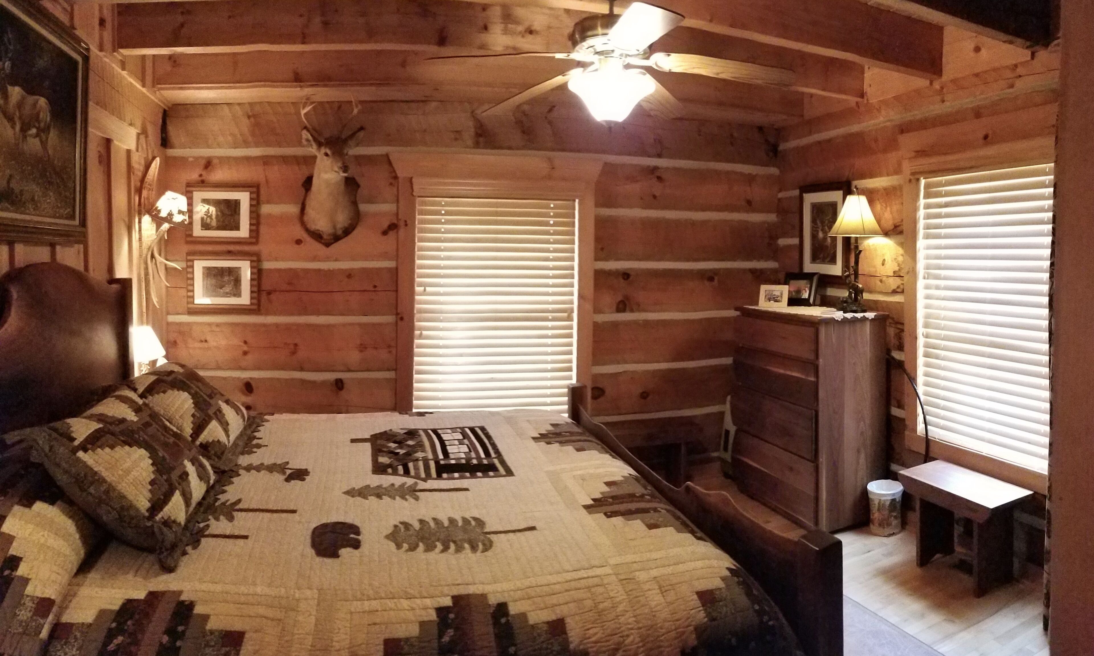 A log cabin bedroom with a bed, a ceiling fan, a mounted deer head, and a dresser.